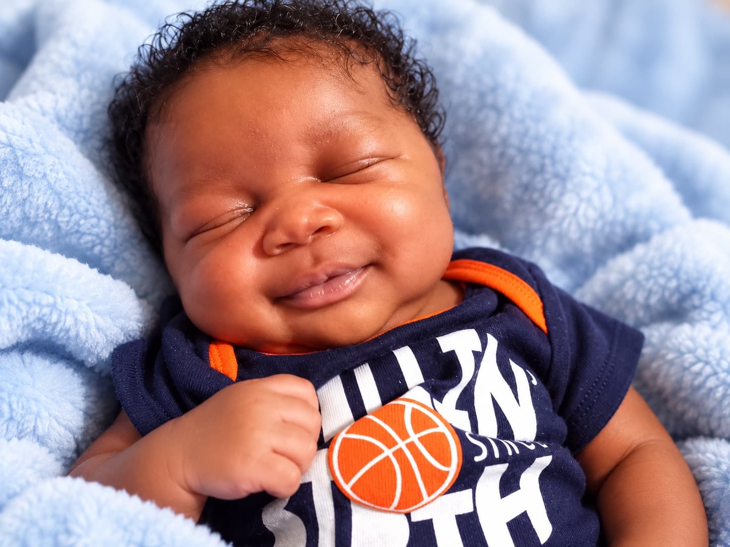 Rheo Moon Moss as a baby, eyes closed, smiling, wrapped in a soft blue blanket.